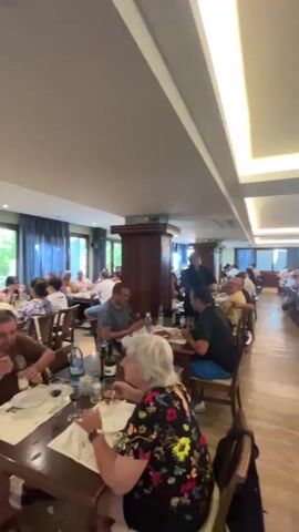 The video shows a busy restaurant interior with many people seated at tables, eating and socializing. The lighting is bright and modern, with long, illuminated ceiling panels. The restaurant appears to be long and narrow, with tables arranged on either side of a central aisle. The decor is simple and functional, with wooden tables and chairs. Several servers can be seen moving between tables, attending to customers. The overall atmosphere is one of a lively and popular dining establishment.