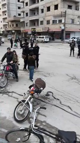 The video depicts a street scene in what appears to be an urban area. Several men in black uniforms and some with tactical gear are present. They stand around motorcycles, a police car, and other vehicles. One motorcycle is lying on its side on the cracked pavement. Some men are standing, while others are walking or mounting motorcycles. Two people are riding on a motorcycle. The buildings in the background are multi-story and appear to be residential or commercial. The overall atmosphere suggests a tense or security-related event.