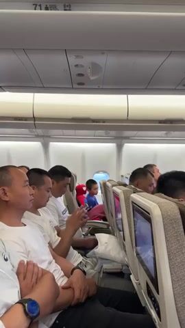 The video shows the interior of an airplane cabin, viewed from the perspective of a seated passenger. Several passengers are visible in their seats. In the foreground on the left, a man wearing a white t-shirt is partially visible, with his arms crossed. Next to him, another man, also in a white t-shirt, is looking at his phone. To his right, a young boy in a blue shirt and red pants is seated by the window, with a large, red stuffed Spider-Man toy next to him. Further down the aisle, other passengers are seated, with some looking at personal devices or the seatback entertainment screens. The overhead compartments and air vents are visible above the passengers. The scene appears to be during a flight, possibly during boarding or a period of waiting, as indicated by the seated passengers and the general ambiance of an aircraft cabin.