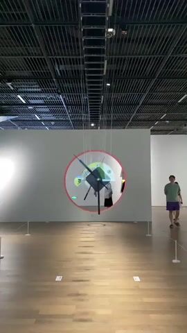 The video shows a modern art gallery. A kinetic sculpture is suspended from the ceiling. The sculpture is made of geometric shapes and appears to be made of metal or plastic. It is rotating, and projected images are displayed on it. A red circle is projected onto the wall behind the sculpture. Two people are visible in the background, walking through the gallery. The floor is made of wood, and the ceiling is a grid of metal panels with lighting fixtures. The walls are white.