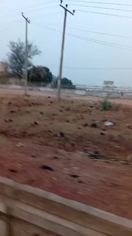 The video depicts a scene of an open field with a reddish-brown, soil-like ground covered with scattered debris, possibly trash or stones. There are a few trees and a building in the background. Utility poles with wires running between them are visible against a pale sky. The camera appears to be moving along a road or path, with a concrete barrier or curb visible in the foreground.