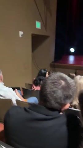 The video shows a dimly lit auditorium filled with a seated audience. The camera appears to be positioned within the audience, looking towards a stage area that is mostly obscured by darkness but has a few lights visible. People are seated in rows, facing the stage. Many individuals are looking at their mobile phones. The overall atmosphere suggests an event is about to begin or is in progress.