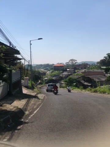 The video captures a view looking down a paved road in a semi-rural or suburban area. The sky is clear and blue, suggesting a sunny day. On the left side of the road, there is a sidewalk and a white fence. Power lines are visible overhead, supported by poles. Several buildings, some appearing to be houses, are scattered along the hillsides on both sides of the road. Trees and green foliage are abundant. A white van and two motorcycles are traveling down the road. One motorcycle is in the foreground, closer to the camera, and another is further ahead. The road curves gently to the right as it descends. The overall impression is of a typical day in a community with a hilly terrain.