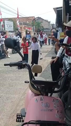A street scene shows people walking and riding motorcycles. In the foreground, a pink scooter is parked. A young girl in a red shirt and blue jeans walks towards the camera, holding a white plastic bag. Next to her, a young girl in a white shirt, black skirt, and a red and white hat walks away from the camera. Other people, some wearing hijabs, walk in the background. The street is lined with buildings and decorated with Indonesian flags. The weather appears to be sunny.