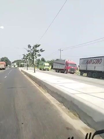 The video shows a view from the driver's seat of a vehicle traveling on a highway. The road is paved and has multiple lanes separated by white lines. To the right of the road, there is a concrete median with a sidewalk and some trees. Several trucks are visible on the road, including a red truck with a gray trailer, a green truck, and a white flatbed truck. The weather appears to be clear and sunny, with a bright sky. The landscape is rural, with trees and vegetation visible on either side of the road. The video is taken from a moving vehicle, indicated by the blurring of the road and surrounding objects.
