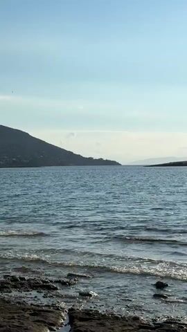 The video shows a static shot of a body of water. The water is calm with small ripples on the surface. Sunlight reflects off the water, creating a shimmering effect. In the background, there are hills and mountains on either side of the body of water. The sky is blue with a few scattered clouds. The foreground shows a rocky shoreline with some seaweed or debris. The overall scene depicts a tranquil natural landscape.