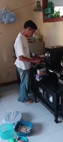 The video shows an indoor scene, possibly a small shop or a home. In the foreground, there are several containers on the floor, including a blue bowl filled with what appears to be peanuts or crackers, a smaller blue container, and a white plastic bag. A young man in a white t-shirt and blue jeans is standing by a black entertainment unit. He is looking down and appears to be interacting with something on top of the unit, possibly a device or electronics.

Further back, on a shelf unit filled with various packaged goods, a young woman wearing a white hijab and a black jacket over a white shirt is interacting with another woman. This second woman is wearing a dark maroon hijab and a matching top with black pants. They seem to be engaged in a transaction, possibly exchanging money or goods. The woman in the maroon hijab is holding what appears to be small packets or bills.

The walls of the room are painted a light yellow, and there are shelves on the wall displaying more packaged goods. There are also two dark chairs visible in the background. The floor is tiled. The lighting suggests an indoor environment, possibly daytime with natural light coming from a window. The overall impression is a casual, everyday scene, potentially involving a purchase or a friendly exchange.