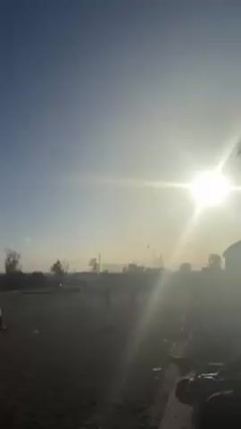 The video shows a dirt field with people running and walking across it. The sun is bright and low in the sky, creating a lot of glare. There are a few trees and some buildings visible in the distance, but they are obscured by the bright sunlight. The camera is initially close to some objects that are mostly in shadow and then pans out to show the field and the people. A plane flies overhead leaving a contrail.