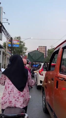The video shows a congested street scene with various vehicles including motorcycles and cars. In the foreground, a person wearing a pink floral shirt and a black hijab is riding a motorcycle. They are facing away from the camera and appear to be stopped in traffic. Beside them, an orange truck is visible. Further ahead, other motorcycles and cars are lined up, suggesting a traffic jam. A green-covered truck is also part of the traffic. In the background, buildings and street signs, including one for "Indomaret," are visible. The sky is overcast, and the overall impression is one of a busy urban environment experiencing traffic congestion.