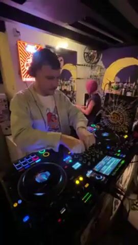 The video shows a man DJing in what appears to be a bar. He is wearing a light-colored shirt over a graphic T-shirt and has headphones on. He is focused on the DJ equipment in front of him, adjusting knobs and using the turntables. Behind him, another person with pink hair is standing at the bar. The bar area is decorated with neon signs, artwork, and bottles of liquor. The lighting is a mix of neon and ambient, creating a lively atmosphere.