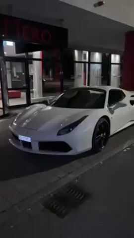 A white Ferrari 488 sits parked in front of a building with the word "RIMERO" in red neon letters over the entrance. The car has black wheels and yellow brake calipers. The building has a modern facade with glass windows and red accent columns. It appears to be nighttime. The pavement is wet. There are no people visible. The camera remains still throughout the video.