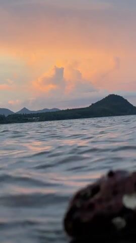 The video shows a low-angle shot of calm water with ripples. In the background, there is a body of water with a distant, tree-covered hill and mountains. The sky above is filled with pink and orange clouds, indicating either sunrise or sunset. In the foreground, a blurry rock is visible.