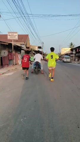 Three young men are walking down a street. One man is on a motorcycle, and the other two are walking beside him. The man on the motorcycle is wearing a white shirt and blue pants. The man to his left is wearing a red shirt with the number 19 on it. The man to his right is wearing a yellow shirt with the number 9 on it. The street is paved and there are buildings on both sides. There are power lines overhead. The sky is clear and blue.