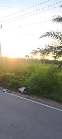 The video opens with a view of a paved road and a grassy field under a sunset sky. A power pole with wires is visible on the left side of the frame. As the video progresses, the camera pans to the right, revealing more of the field and the setting sun. The sky is filled with hues of orange, yellow, and pink. The scene is dominated by natural elements, with tall grass and some scattered trees forming a silhouette against the vibrant sky. There are no people or animals visible in the video. The overall atmosphere is serene and tranquil, capturing the beauty of a rural sunset.