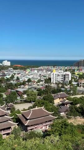 This video shows an aerial view of a coastal town with a clear blue sky. In the foreground, there are traditional buildings with layered, ornate roofs, surrounded by lush green trees. These structures appear to be part of a temple complex or similar cultural site. Beyond this, the town unfolds with a mix of smaller houses and more modern buildings, including some taller white structures that could be hotels or apartment buildings. The town stretches towards the sea, where the blue ocean meets the horizon. In the distance, faint outlines of hills or mountains can be seen. The overall scene suggests a warm, sunny day in a picturesque location, likely a tourist destination in a Southeast Asian country, given the architectural style. There are no people or active movements visible in the frame.