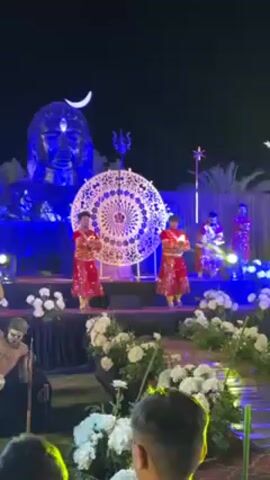 A nighttime stage scene is in view, featuring several performers. In the foreground, two figures, clad in traditional red costumes and holding lit candles, stand at the front of the stage. Behind them, centered, is a large decorative circular piece of art. Further back on the stage, additional performers in similar red costumes are positioned. Behind the performers is a giant sculpture of a face lit with blue light, and a large crescent moon visible in the dark sky behind it. The stage is decorated with white flowers and lit with blue lights. In the lower left corner of the frame, a partly visible figure in a costume stands to the side. In front of the stage is a blurred audience member.