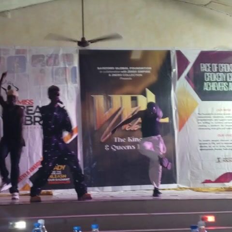 Four men are performing on an indoor stage, likely at an event or competition. The stage is set with two large vertical banners in the background.

The performers are dressed in various outfits:
- One man in a black tank top and light-colored pants is positioned towards the upper right, striking a pose with one leg extended and leaning back.
- Another man in a black outfit adorned with white spots or patterns, wearing a black cap, is centrally located, mid-pose with arms extended outwards.
- A third man in a black long-sleeved shirt, black pants, and a light-colored cap is in the lower-left portion of the frame, also striking a dynamic pose.
- The fourth man, wearing a dark, possibly camouflage or tactical-style outfit, is at the very bottom of the frame, slightly crouched.

In the background, the top banner features a dark brown and gold design with text that reads: "SAMZORO GLOBAL FOUNDATION In collaboration with ZIIGII EMPIRE & 2NERO COLLECTION Presents" and prominently displays "Vibez The Kings & Queens Event". The bottom banner is lighter in color, possibly white or cream, with text visible including "MISS BEAUTY & BRAINS" and "UNLEASH YOUR RADIANT PERSONALITY".

To the left, a white ceiling fan with brown blades is visible hanging from the ceiling. On the right side of the stage, several plastic bottles of water and soft drinks are arranged on a table, with a partially visible sign that reads "VVIP". The stage appears well-lit, suggesting a formal performance setting. The men's poses indicate they are engaged in a choreographed routine or a talent showcase.