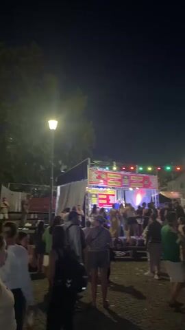 The video shows a crowded outdoor event at night. In the foreground, several individuals are visible from behind, looking towards a stage area. The stage is illuminated with colorful lights and has a banner across the top. Behind the stage, more people are gathered, and tents or stalls are set up. Festive string lights are hung across the scene, casting colored lights against the dark sky. The ground appears to be paved. People are milling about, suggesting a lively atmosphere, possibly a festival or a street party.
