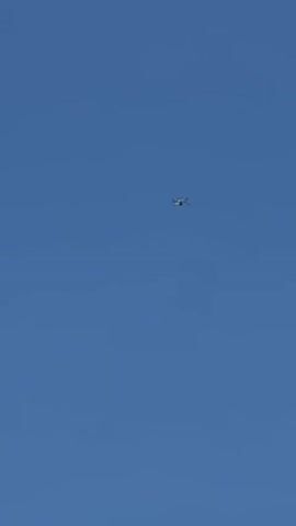 A white quadcopter drone with four visible propellers flies and hovers in a clear, bright blue sky. The drone maintains a relatively stable position, appearing to move slightly closer and then further from the camera while remaining largely centered in the frame. No other objects, people, or environmental features are visible.