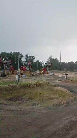 The video shows an outdoor scene with several oil pumpjacks in the background. The sky is overcast and appears to be gray. The ground in the foreground is dirt with some patches of grass. There is a dirt road or path winding through the scene. The oil pumpjacks are red and appear to be operational. Trees are visible in the background, forming a line of forest. There are also utility poles or similar structures scattered throughout the area. The overall impression is of an industrial oil field setting under a cloudy sky.