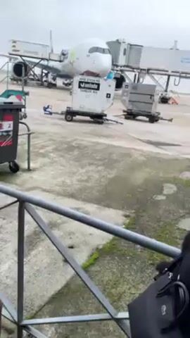 The video shows an airplane parked at a terminal, with ground support equipment nearby. The plane has a white body with colorful accents, and is connected to the terminal via a jet bridge. In the foreground, there's a metal railing, a trash can, and a person partially visible on the right side of the frame. Two white cargo containers on small trailers are positioned on the concrete ground between the railing and the airplane. The ground is cracked and stained. The sky appears overcast. The text on the cargo containers reads "unilode" and "TARE 58 KG".