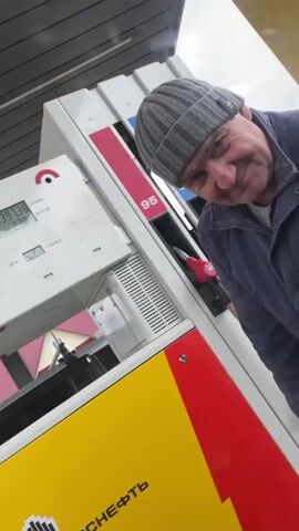 A man wearing a grey knit cap and a blue jacket stands by a ROSNEFT gas pump. The pump is painted yellow, red, and white. The pump display shows 140.76 rubles and 2.226 liters. The man is looking at the camera and smiling slightly. He then turns away and appears to be putting the gas nozzle into a car.
