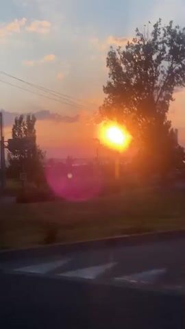 The video shows a view from a vehicle moving along a road during sunset. The sun is visible, casting a bright orange glow and creating lens flare. There are trees and utility poles on either side of the road. Some clouds are present in the sky, reflecting the sunset colors. Road signs and streetlights are also visible. The environment appears to be rural or suburban. The movement of the vehicle causes the scene to shift and blur, particularly the foreground.