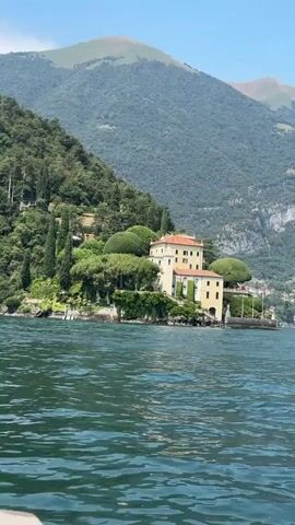 The video shows a serene lake with gentle waves lapping against the shore. A grand, pale yellow villa with red-tiled roofs sits majestically on a hill overlooking the water. Lush green trees and manicured hedges surround the villa, with tall, slender cypress trees adding a touch of elegance. Behind the villa, a verdant mountainside rises, covered in dense foliage, reaching towards a clear blue sky. The scene evokes a sense of tranquility and timeless beauty.