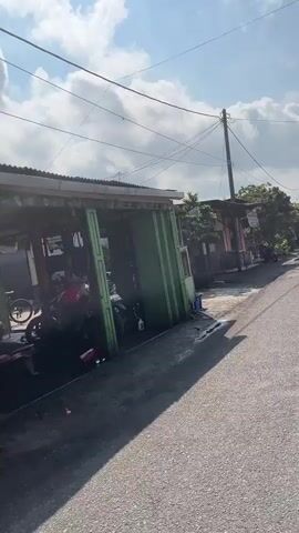 The video shows a street scene with buildings and vehicles. On the left, there is a covered structure with green metal posts and a corrugated metal roof. Several motorcycles are parked inside and around this structure. The ground in front of the structure is wet and appears to be asphalt. A few people can be seen in the background, but their faces are not clear. To the right of the structure, the street continues, with more buildings lining the other side. Overhead power lines are visible, and the sky is partly cloudy. The overall impression is of a street in a residential or commercial area, possibly in a tropical climate given the sunlight and vegetation.