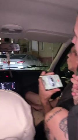 The video shows three young men in the front seats of a car at night. The person in the foreground on the right is out of focus, with their head and shoulder visible. The person in the middle is in focus, sitting in the driver's seat, holding a smartphone horizontally. The phone screen displays a call interface with a large gray circle with two dots, resembling eyes, and what appears to be a microphone icon. The person in the middle is looking at the phone and speaking. They have tattoos on their arm and leg. The person on the left is mostly out of focus and their back is to the camera. In the background, through the car's windshield, several cars are parked in front of a building. The overall scene suggests a casual car ride at night, with the occupants engaged in a phone call.