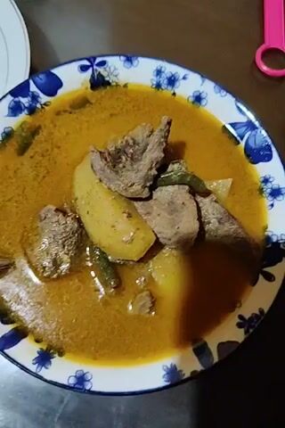 The video shows a blue and white patterned plate filled with a yellow-orange stew containing potatoes, meat, and green beans. The camera moves slightly around the plate, showing the food from different angles. Other objects visible in the background include a white plate, a pink object, and a wooden table.