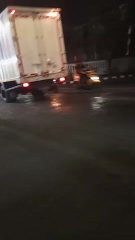 The video shows a street scene at night. A large white truck is on the left side of the road. Several motorcycles are also on the road, with headlights visible. Some streetlights are on, illuminating the wet asphalt. There are buildings and trees visible in the background, along with a white fence. One shot shows a person standing near a food stall. The footage is blurry and appears to be taken from a moving vehicle.