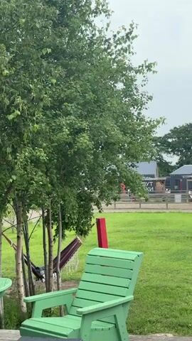 The video opens on a lush green lawn with a large tree on the left. A red and white striped hammock is visible hanging from the tree branches, along with a bright green Adirondack chair in the foreground. In the background, several small red sheds with star decorations are lined up. The scene then shifts to a wide shot of a grassy field. Two people, one wearing a long dark coat and the other a white t-shirt and dark shorts, walk across the field, followed by a small tan dog. A yellow building and several hay bales are visible in the distance. The video concludes with a view of rolling hills and a body of water in the background.