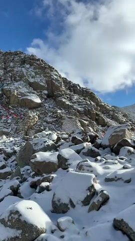 The video showcases a vast, snow-covered mountainous landscape under a bright blue sky dotted with fluffy white clouds. The ground is largely composed of large, grey boulders and rocks, all dusted with a fresh layer of white snow. In the background, majestic, rugged mountains, also partially covered in snow, rise against the horizon. Sunlight occasionally breaks through the clouds, illuminating the snowy peaks and casting shadows across the terrain.

Scattered throughout the rocky landscape are several cairns, which are small stacks of stones. At one point, a person is seen walking away from the camera, ascending a snowy path through the rocks. This individual is wearing a distinctive lime green hooded jacket, black pants, and is using trekking poles. They also have a black shoulder bag. The overall scene depicts a remote, high-altitude environment, possibly a mountain pass or a hiking trail, with clear and cold weather conditions.