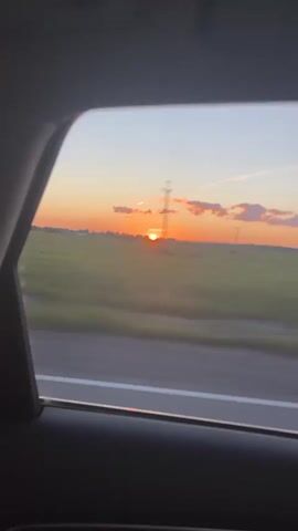 The video captures a sunset viewed from inside a vehicle. The sun is low on the horizon, casting an orange glow across the sky and illuminating scattered clouds. A field of green crops stretches across the landscape, with a road visible in the foreground. Power lines and distant structures can be seen on the horizon. The viewpoint is through a car window, with the interior of the vehicle, specifically the dark gray frame of the window and part of a dark interior panel, visible in the foreground. The overall impression is one of movement, as the landscape appears slightly blurred, suggesting the vehicle is in motion.