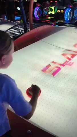 The video shows two young children playing air hockey in an arcade. The child in the foreground, on the left, is wearing a light blue shirt and leopard print shorts and is using a red paddle. The child in the background, on the right, is wearing a green t-shirt. The air hockey table has a bright, illuminated surface with a red and clear plastic railing. The background shows other arcade games with flashing lights and screens. The children are actively engaged in playing the game, with their paddles in motion.