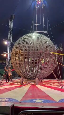 The video shows a large metal sphere at a circus. The sphere, made of a crisscross pattern of metal bars, is supported by a frame on a stage. Two men stand near the base of the frame. The stage is red and white striped. A spotlight shines on the sphere from above. In the background, an empty, green-colored seating area is visible. The top of a circus tent is visible, with rigging and lighting attached.