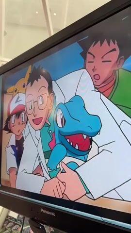 The video displays a television screen showing an animated scene from the Pokémon anime. The first frame shows a close-up of Professor Oak from the shoulders up. He has gray hair, dark eyebrows, and is wearing a red collared shirt under a white lab coat. He has a slight smile on his face and is looking slightly to his left. Behind him, a blue and teal machine with a pink knob is visible. The subsequent frames show a wider view of the scene. Professor Oak is now holding a blue Pokémon with red and white accents. To his left, Ash Ketchum is visible wearing his signature red and white hat and blue vest. Pikachu is partially visible behind Ash's hat. To Professor Oak's right, Brock is leaning in, looking surprised. The environment appears to be an indoor setting, possibly a research lab or a conference room, suggested by the clean lines and the presence of the television itself in the foreground, which has a Panasonic logo at the bottom. The camera is positioned at an angle, capturing the television screen from a slightly elevated perspective.