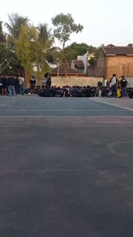 The video shows a group of young men sitting on the ground in a large outdoor area, possibly a sports court or a schoolyard. Some individuals are standing and walking around. In the foreground, two people are walking towards the left of the frame. One of them is wearing an orange jumpsuit and a white mask with what appears to be bunny ears. The background includes buildings, trees, and a cell tower. The overall atmosphere seems informal.