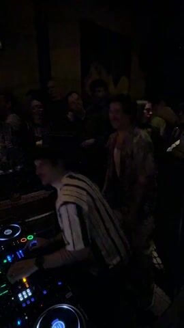 The video depicts a DJ in a striped shirt working on a mixing console in a dark club. A small crowd of people are standing around him, some are dancing and raising their arms. The DJ looks focused and is working the equipment. The room appears to be dimly lit with a few colored lights.