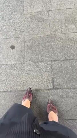 The video shows a first-person perspective of someone standing on a stone-tiled floor. The person is wearing dark-colored pants with a pinstripe pattern and transparent, pointed-toe heels in a muted pink or purple hue. The camera is pointed downwards, focusing on the shoes and the lower part of the pants. The lighting suggests it is daytime. There is a small, dark stain visible on one of the stone tiles. The person appears to be stationary at the beginning of the clip and then takes a step forward, causing a slight shift in the perspective and movement of the shoes.