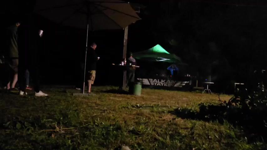At night, a group of people are gathered outside under a large umbrella. A DJ is visible under a green tent, with a "BALKAN STATION" banner behind them. The ground is covered in grass and some debris. The lighting is dim, with some spotlights illuminating the DJ booth.