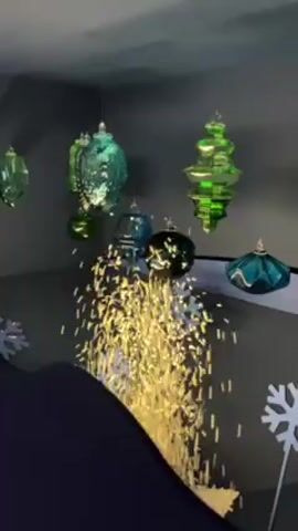 The video displays a dark room adorned with green and blue Christmas ornaments suspended from the ceiling and white snowflakes on the floor. A stream of gold glitter rains down from an unseen source, filling the lower portion of the frame. A dark blue object, presumably furniture, sits at the bottom, partially visible. The overall aesthetic suggests a holiday-themed display or decoration.