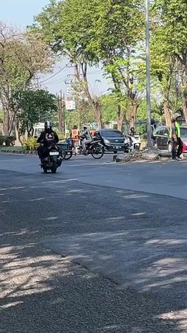 The media file captures a street scene in Ngampelsari, Indonesia, during daylight. The subjects are people riding motorcycles and what appears to be a cart being pulled by a motorcycle. Several motorcycles are visible on the asphalt road. In the background, there are trees lining the street, suggesting an outdoor, possibly urban or suburban, setting. Some parked cars are also present, including a black SUV and a lighter-colored sedan. A person in a bright green vest stands near one of the parked vehicles. The lighting indicates it is likely daytime, with sunlight casting shadows. The overall mood appears neutral, depicting everyday street activity. No visible text is present in the media.