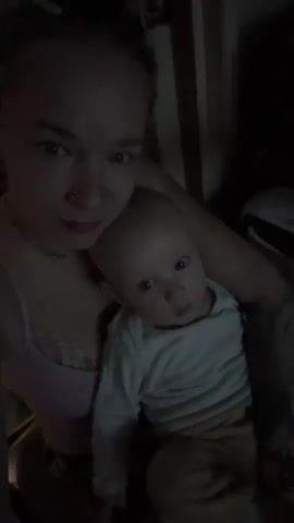 A woman and a baby are seen in a dimly lit room. The woman is holding the baby in her arms. In the background, another person appears to be lying down. The room contains furniture, possibly a bed or couch, and a projector screen displaying an image. The overall scene appears to be a casual, indoor setting.