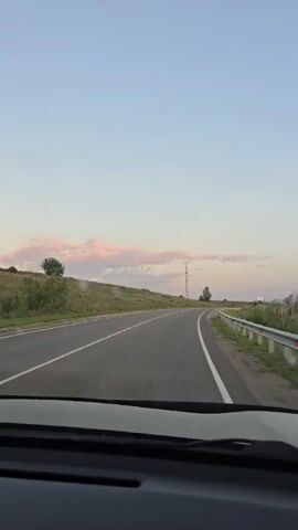 The video captures a first-person perspective from inside a vehicle traveling on a paved road. The road is marked with white lines, indicating two lanes. The landscape outside the vehicle consists of rolling hills covered in dry grass and scattered trees. In the distance, there are power lines and a tall tower. The sky is clear and pale blue, with a few wispy clouds tinged with pink, suggesting it might be dawn or dusk. The vehicle's dashboard is partially visible in the foreground. The video appears to be a short clip of a continuous drive through a rural area.