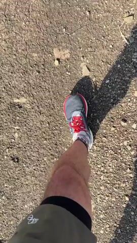 A first-person perspective video shows someone walking on dry, cracked ground. The person is wearing dark green shorts with black trim, grey socks, and athletic shoes with red laces and a navy blue and white design. A shadow of the person's legs and upper body is cast on the ground. The camera moves with the person's steps, capturing the movement of their feet and legs. The environment appears to be outdoors on a sunny day.