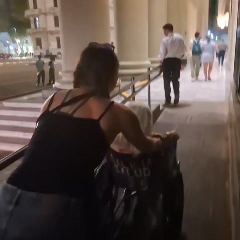 A woman pushes a person in a wheelchair in front of a building with many large columns. The person in the wheelchair has white hair and the wheelchair has "GALICIA SALUD 25" on the back. Several people are walking near the columns, including a man in a white shirt. The woman pushing the wheelchair is wearing a black tank top and jeans. There is a crosswalk in the background.