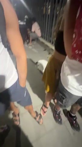 The video shows a close-up of a man with curly dark hair and tattoos on his arms, wearing a white t-shirt with red bandana print on the sleeves. He is looking directly at the camera with his arms crossed. Behind him, a woman with blonde wavy hair is partially visible, wearing a yellow skirt. Other people are in the background, out of focus, in what appears to be an outdoor setting at night, with some artificial lighting. There is a metal fence in the background, and some dark objects that could be chairs or tables.