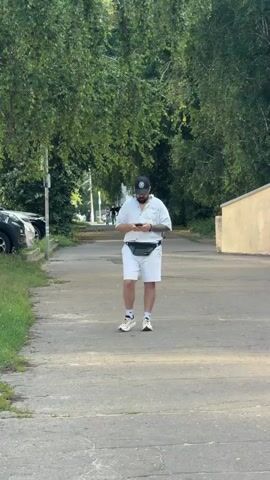 A man walks down a paved path lined with trees on either side. He is wearing a white short-sleeved button-down shirt, white shorts, a black baseball cap, and white sneakers. He has a black fanny pack around his waist and is looking down at his phone. To his left are parked cars and grass, and to his right is a beige building. The scene appears to be set in a park or residential area, with lush green trees suggesting a warm, perhaps summer, day. The lighting indicates it is daytime, likely late afternoon due to the angle of the shadows. The man's focus on his phone suggests he is engaged in a digital activity. The overall atmosphere is casual and everyday.